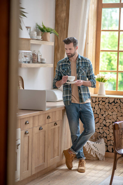 Mann mit Tasse in der Hand steht in der Küche und liest Nachrichten auf dem Laptop - Foto, Bild