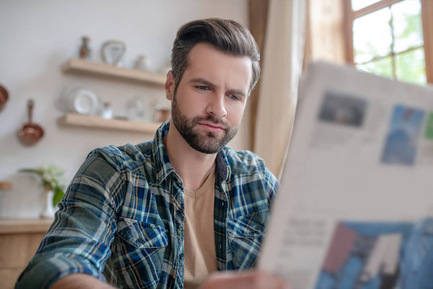 Man in a checkered shirt sitting at the table, reading newspaper and looking serious - Foto, imagen