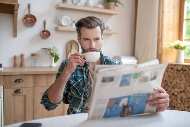Homme en chemise à carreaux assis à la table, prenant un café et lisant un journal - Photo, image