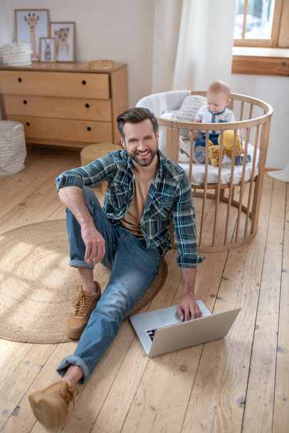 Dark-haired father working on a laptop while his little son playing in a playpen - Foto, afbeelding