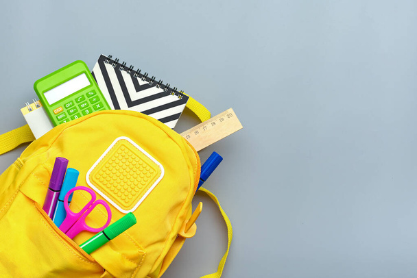 Back to school, education concept. Yellow backpack with school supplies - notebook, pens, ruler, calculator, scissors isolated on gray background. Top view. Copy space Flat lay composition Banner. - Фото, изображение
