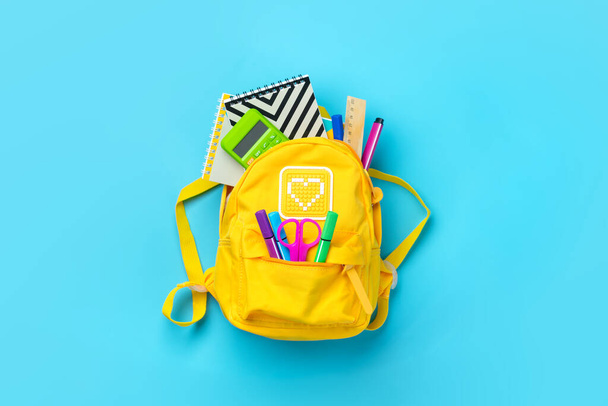 Back to school, education concept. Yellow backpack with school supplies - notebook, pens, ruler, calculator, scissors isolated on blue background. Top view. Copy space Flat lay composition Banner. - Photo, Image
