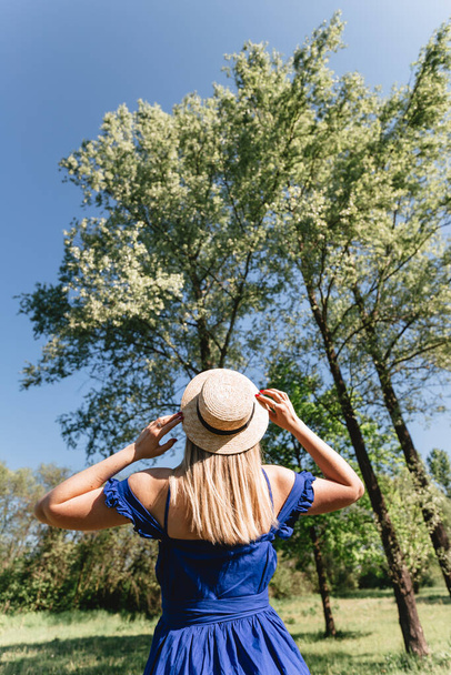 Egy szalmakalapot és kék ruhát viselő fiatal nő portréja, aki a vidéki tájban áll. Nyári időszámítás, aktív életmód. - Fotó, kép