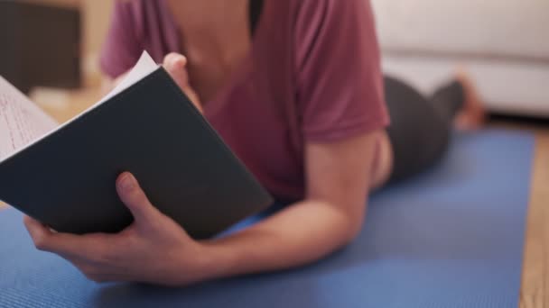 Fiatal, aktív nő sportruhában irodalmi könyvet olvas, miközben edzés után jóga matton fekszik, otthon pihentető szabadidős tevékenység, egészségügyi és oktatási hobbi, elszigetelt a kovid világjárvány idején - Felvétel, videó