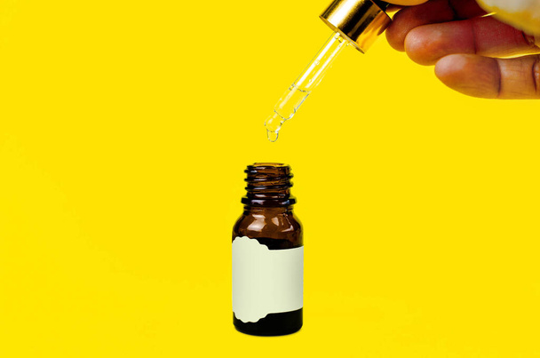 Little bottle of essential aroma oil with pipette and drops in hand on yellow background - Photo, Image