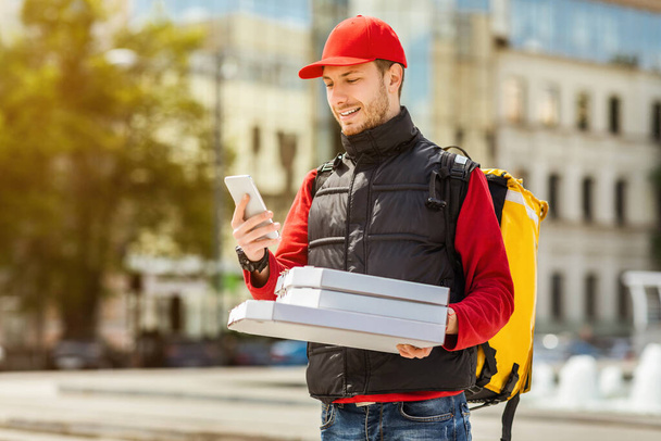 Mensajero usando teléfono entregando pizza y comida de restaurante caminando al aire libre
 - Foto, Imagen