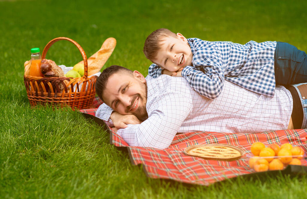 glimlachende vader met zijn kleine zoon liggend op geruite deken samen tijdens de picknick - Foto, afbeelding
