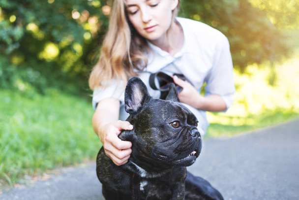 Beautiful young woman playing with her French bulldog in a park outdoors. Close-up portrait of pleased dog with short black coat. Soft focus on a contented dog. - Photo, image