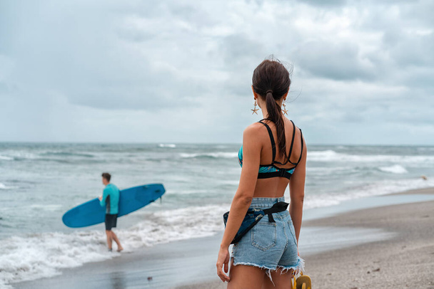 Καλοκαίρι ενεργό. Μια γυναίκα βλέπει έναν άντρα να περπατάει με μια σανίδα του σερφ στη θάλασσα. Πίσω όψη. - Φωτογραφία, εικόνα