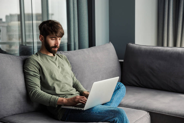 Image of focused young man in casual wear using laptop while sitting on sofa in living room - Foto, immagini