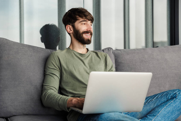Image of joyful young man in casual wear using laptop and smiling while sitting on sofa in living room - Foto, Bild