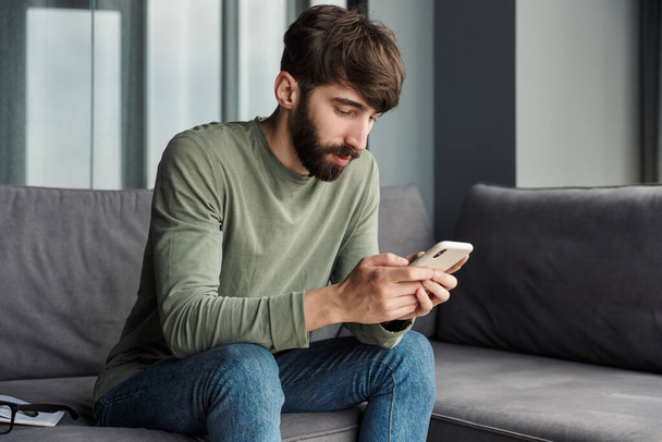 Image of handsome young man typing on mobile phone while sitting on sofa in living room - Fotoğraf, Görsel