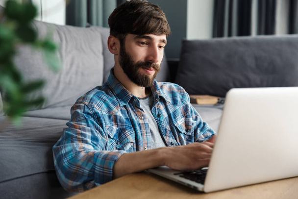 Image of pleased bearded man working with notebook while sitting on floor in living room - Foto, afbeelding