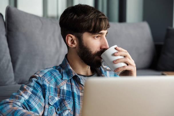 Image of handsome bearded man drinking coffee and using notebook while sitting on floor in living room - Foto, immagini