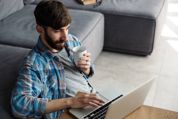 Image of handsome bearded man drinking coffee and using notebook while sitting on floor in living room - Foto, imagen