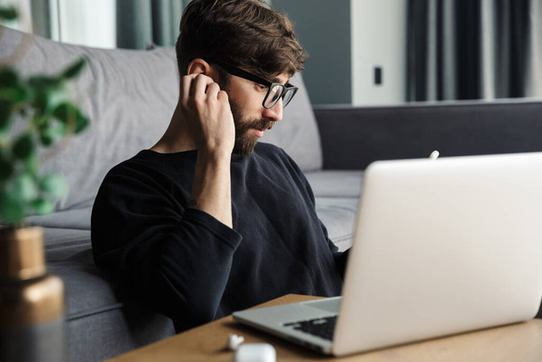 Image of serious man in eyeglasses using notebook and wireless earphones while sitting in living room - Foto, Imagem