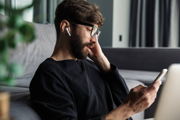 Image of young man in eyeglasses using cellphone and wireless earphones while sitting in living room - Foto, Imagen