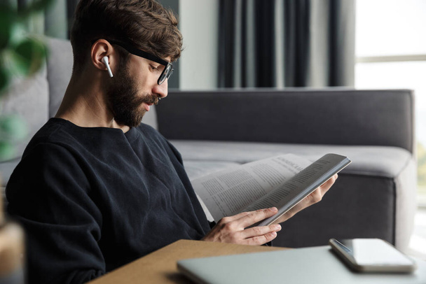 Image of focused man in wireless earphones drinking coffee and reading magazine while sitting in living room - Foto, imagen