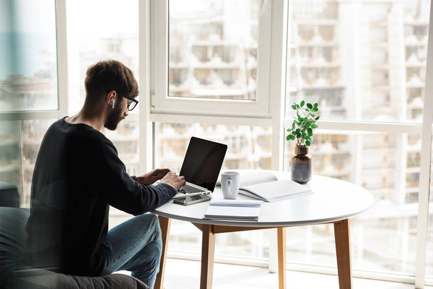 Image of young serious man using wireless earphones and working with laptop while sitting at table in living room - Photo, Image