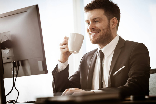Business concept. Successful young businessman at work. Manager sitting at the office table happy drinking coffee from cup. Man smiling in suit indoors on glass window background. - Foto, immagini