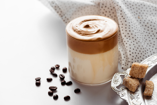 delicioso café Dalgona em vidro perto de grãos de café, açúcar mascavo na bandeja de prata e guardanapo no fundo branco
 - Foto, Imagem