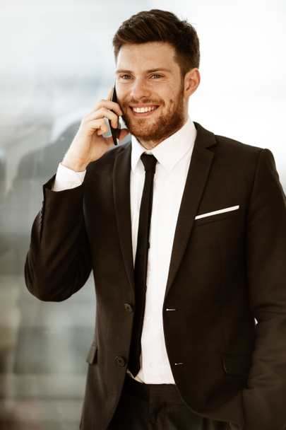 Business concept. Happy smiling young businessman standing in office talking on a cell phone getting good news about his work. Man in suit indoors on glass window background. - Photo, Image