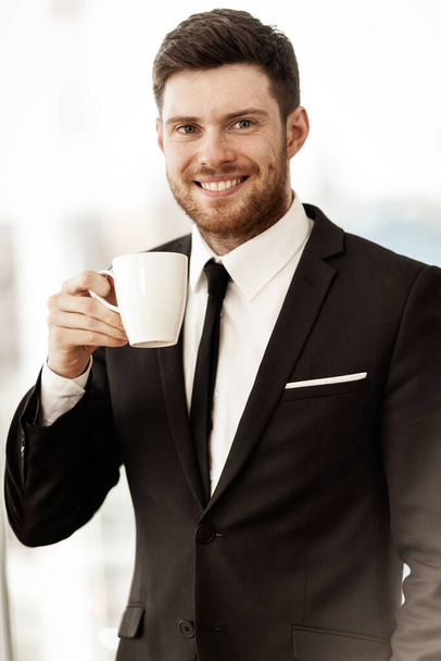 Bedrijfsconcept. Succesvolle jonge zakenman aan het werk. Manager staat op kantoor en drinkt graag koffie uit de beker. Man glimlachend in pak binnen op glazen raamachtergrond. - Foto, afbeelding