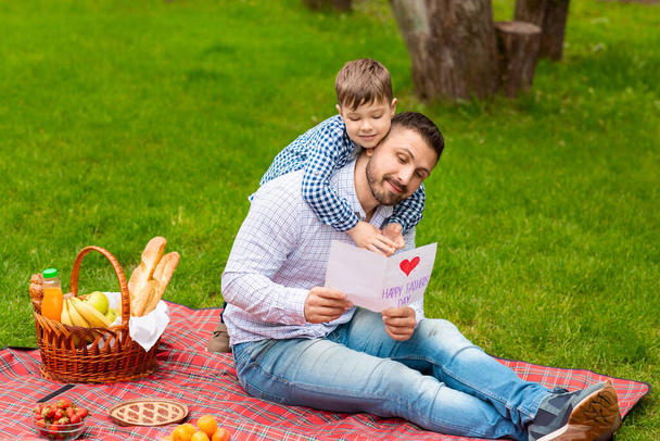 Překvapení pro tatínka. Usmívající se táta čtení otců den ručně vyrobené karty od svého syna při pikniku v lese - Fotografie, Obrázek