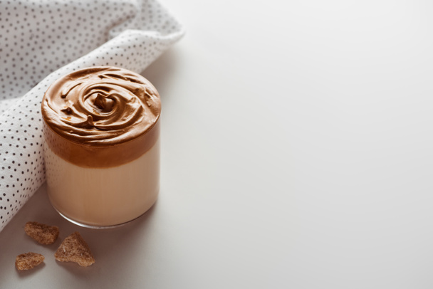 heerlijke Dalgona koffie in glas in de buurt van servet en bruine suiker op witte achtergrond - Foto, afbeelding