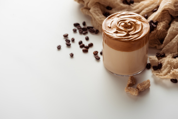 delicious Dalgona coffee in glass near coffee beans, cloth and granulated brown sugar on white background - Foto, immagini