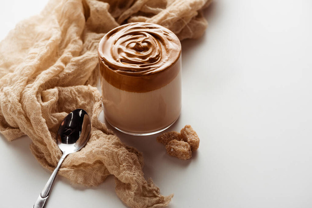 вкусный кофе Dalgona в стакане возле ложки, ткани и гранулированного коричневого сахара на белом фоне
 - Фото, изображение