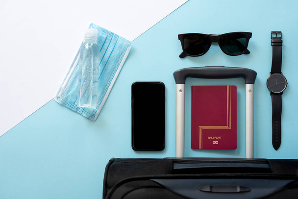 Horizontal color image with an overhead view of a travelers accessories on a 2020 summer holidays with the coronavirus on a blue and white background. Black suitcase, face mask, hydroalcoholic gel, clock, smartphone, sunglasses and passport. - Фото, зображення
