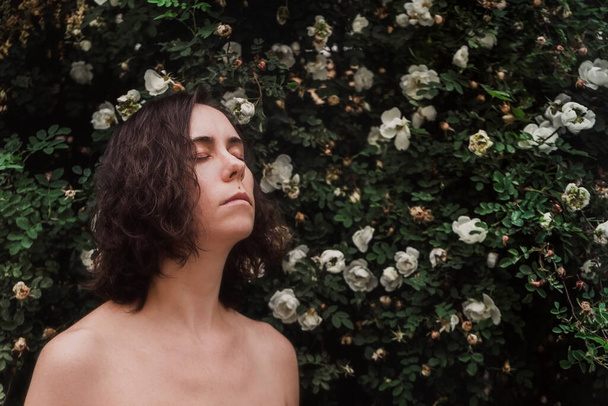 Ein Mädchen mit geschlossenen Augen und nackten Schultern inhaliert den Duft von Gartenrosen. Meditation vor einer lebendigen grünen Wand mit Blumen - Foto, Bild