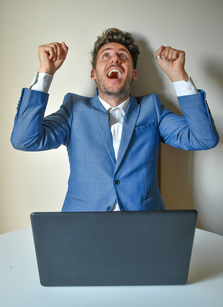 Uomo in giacca e cravatta seduto davanti a un computer, che alza le braccia per la felicità, il successo - Foto, immagini