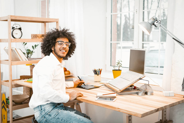 Intelligent freelanser smiles at camera sitting at home workplace. Creative young businessman working from home. Distant work or remote work concept. Toned image - Foto, imagen