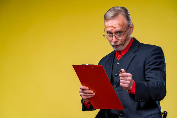 Senior businessman sitting with folder over yellow background in suit looking at camera in red shirt . Business concept - Foto, Bild