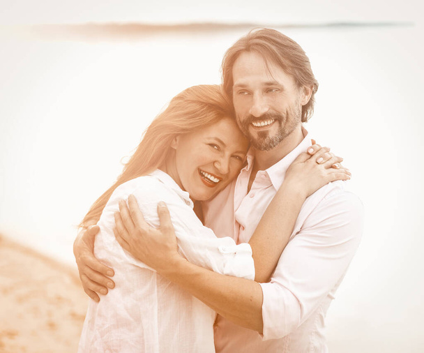 Beautiful couple stand embracing against sea or ocean background. Smiling man and woman in white clothes hugging each other. Tinted image - Фото, изображение