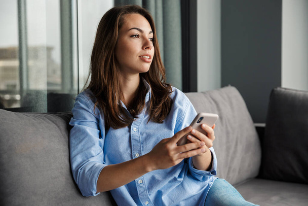Image of caucasian young focused woman using mobile phone while sitting on sofa at living room - Foto, Imagem