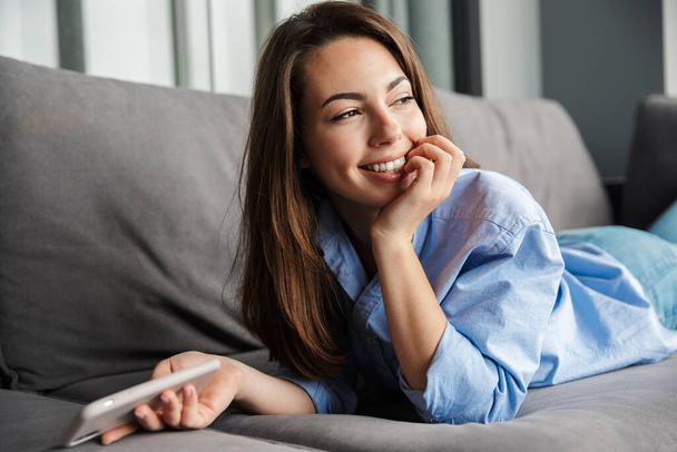 Image of young joyful woman smiling and using mobile phone while lying on sofa at living room - Fotoğraf, Görsel