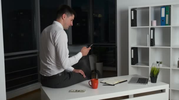 Handsome man linger at work until late at night sitting at desk in office with panoramic windows and holding phone - Video