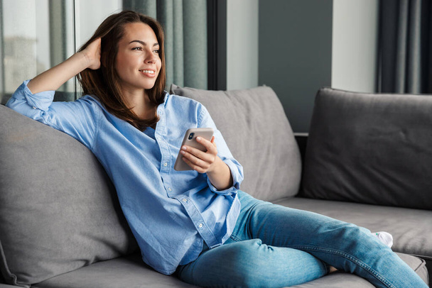 Image of beautiful joyful woman smiling and using mobile phone while sitting on sofa at living room - Foto, afbeelding