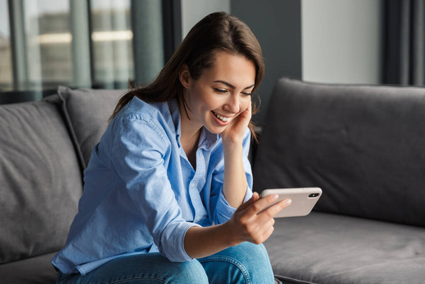Image of beautiful cheerful nice woman smiling and using mobile phone while sitting on couch at living room - Фото, зображення
