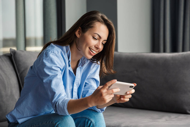 Image of beautiful cheerful woman playing video game on mobile phone while sitting at living room - Фото, изображение