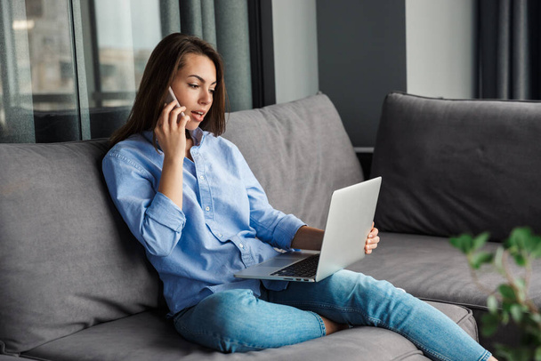 Image of young focused woman talking on mobile phone and using laptop while sitting at living room - Photo, image