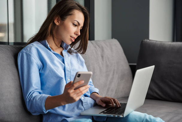 Image of young focused woman working with laptop and cellphone while sitting on couch at living room - Photo, Image