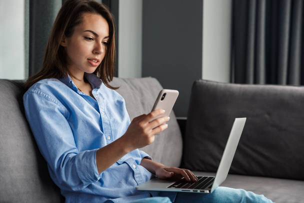 Image of young focused woman working with laptop and cellphone while sitting on couch at living room - Fotografie, Obrázek