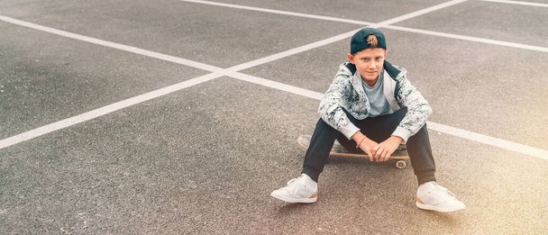 Teenager skateboarder boy with a skateboard sitting on asphalt playground and resting after he made tricks. Youth generation Freetime spending concept image. - Photo, image