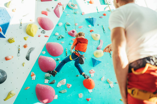 Father and teenager son at indoor climbing wall hall. Boy is hanging on the rope using a climbing harness and daddy belaying him on the floor using a belay device. Happy parenting concept image. - Фото, изображение
