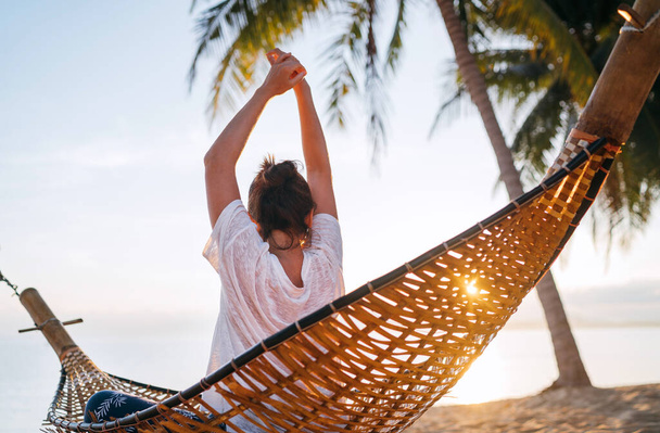 Young woman awaking and meeting morning sunrise sunlight sitting in a hammock and lazy stretching her body raising arms up on the sandy beach under the palm trees. Calm exotic places vacation concept - Fotoğraf, Görsel