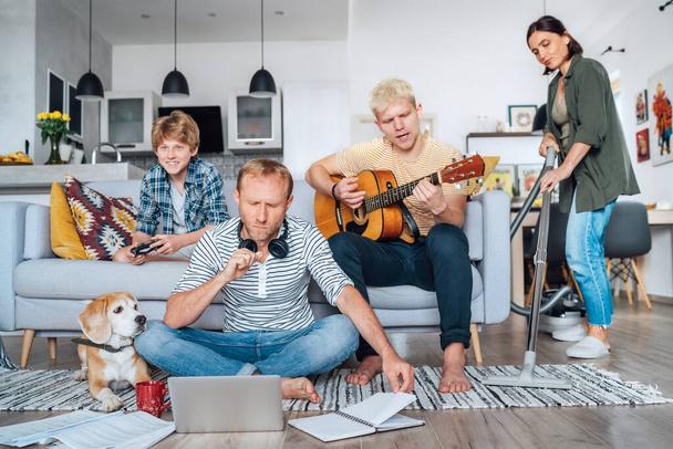 Quarantine time: Family at living room together. Father distance working using laptop, mother cleaning the room, older son playing acoustic guitar and singing a songs, little son playing video console - Фото, зображення